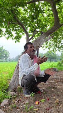 Senior indian farmer sitting under tree rubbing and consuming tobacco khaini in rural field