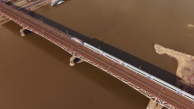Aerial Freight Train Crossing Bridge Warszawa At Dawn, Muddy Vistula Below, Rustred Steel Girders, Solitary Cargo Wagons Moving Slowly, Industrial Atmosphere And Quiet Urban Riverbank.