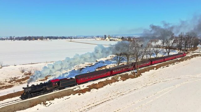 A steam train moves along a track in a snowy area. The train billows smoke as it passes fields covered in white snow. The sun shines brightly, highlighting the winter scenery.