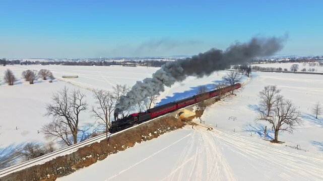 A steam train moves along a track in a snowy area. Smoke rises from the train as it rolls past bare trees. The sun shines in the clear blue sky. The landscape stretches far in all directions.