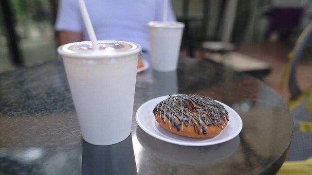 Iced Coffee And Chocolate Donut On Table, Outdoor Cafe Scene With White Disposable Cup And Straw, Chocolatedrizzled Donut On Plate, Reflective Metal Table, Blurred Seated Customer