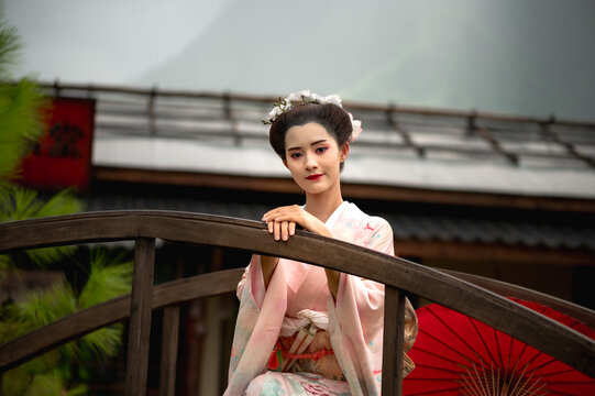 Graceful Japanese geisha wearing national traditional kimono in sweet pastel floral pattern and obi sash, standing on a traditional wooden bridge