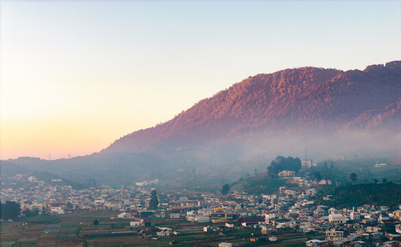 Aerial view of Dieng Plateau with a town nestled in the valley, surrounded by misty mountains under the soft light of golden hour in Indonesia.