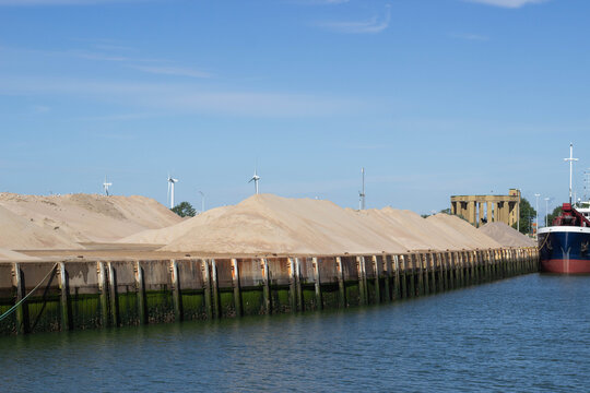 Large sand piles along a waterfront dock with a cargo ship moored nearby and wind turbines visible in the background under a clear blue sky