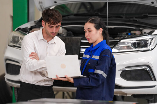 Car mechanic customer laptop garage repair, female mechanic discussing vehicle battery service with male customer beside open hood in auto workshop
