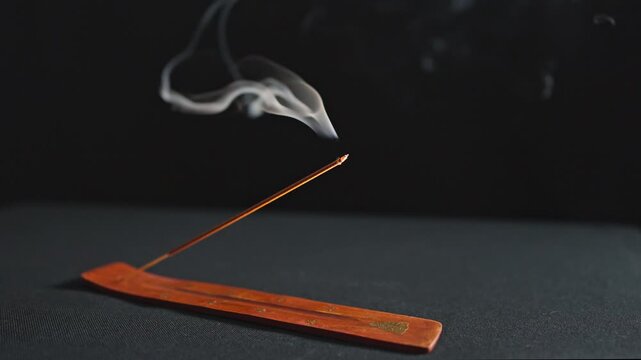 A single incense stick smolders and emits white smoke against a dark background in slow motion clip.