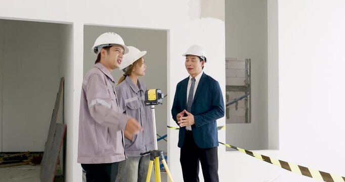 Asian female engineer points toward unfinished wall while male contractor and manager discuss construction site safety inspection, project coordination, progress tracking, work direction today.