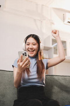 Beautiful young asian woman sitting on sofa and holding joystick playing online video game in living room at house