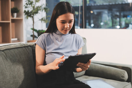 Freelance Asian woman holding mobile phone typing on laptop and working from home. Happy young girl sitting on sofa in living room.