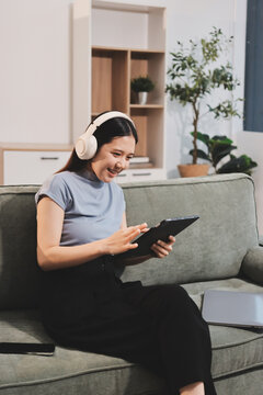 Freelance Asian woman holding mobile phone typing on laptop and working from home. Happy young girl sitting on sofa in living room.
