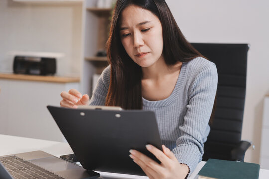 Young Asian woman sits at office desk rubbings her eyes and temples feeling pain. Stress. And exhaustion from long hours working on the computer