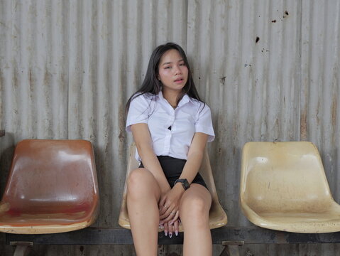 A young Asian woman with long straight black hair, wearing a white short-sleeved collared shirt and black skirt, sits on a worn beige plastic chair against a rusty corrugated metal wall