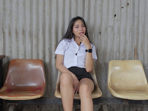 A young Asian woman with long straight black hair, wearing a white short-sleeved collared shirt and black skirt, sits on a worn beige plastic chair against a rusty corrugated metal wall