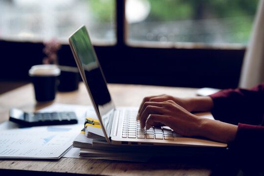 Business professional working on a laptop with financial charts, calculator, documents and coffee on a wooden desk, representing finance, accounting and office work.