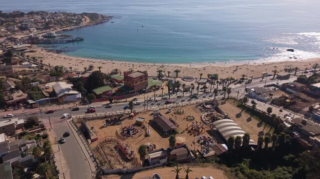 Approaching shot over quiet residential streets and amusement park revealing the crescent shaped bay, sandy beach and calm turquoise waters. El Quisco, Chile