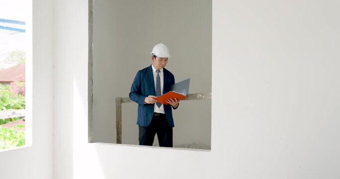 Asian male engineer reviews red project folder, looks across unfinished building room, checks construction progress, site safety compliance, wall condition, and document report indoors today.