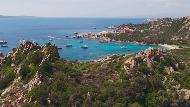 Epic aerial flying over granite cliffs to reveal luxury sailing yachts in turquoise blue water of a natural protected harbor on the Isola di Spargi in the Maddalena Archipelago in Sardinia, Italy.