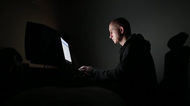 Hacker programming and typing on laptop computer at night his face illuminated by the screen in a dark clandestine room