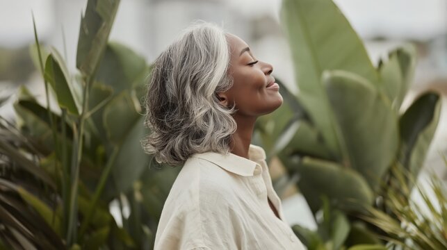 A portrait of a woman in her early forties with natural grey hair standing among large green tropical leaves with eyes closed and a gentle smile enjoying fresh air in soft daylight
