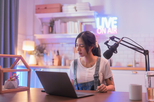Focused young Asian woman concentrating on her laptop while working from a home podcast studio, with a glowing "Live On Air" neon sign and microphone in the background.