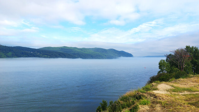Misty morning view where Angara River meets Lake Baikal, fog hovering over calm blue water with green forested hills in background, dirt path along grassy shoreline, serene Siberian landscape