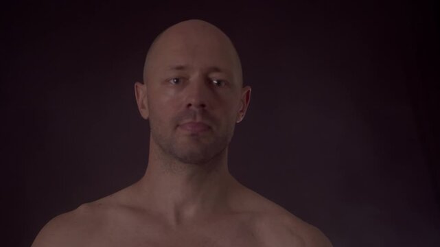 Focused shirtless man juggling tennis balls in dark studio. Caucasian male with bald head demonstrating hand-eye coordination, focus, and dexterity against dark background.
