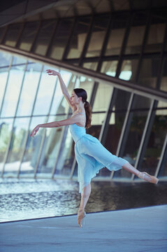 Professional female ballet dancer performing an elegant arabesque on pointe beside reflective water and contemporary architecture.