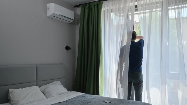 Man approaches the window, closes it, and turns on the air conditioner with a remote control to cool down from the summer heat