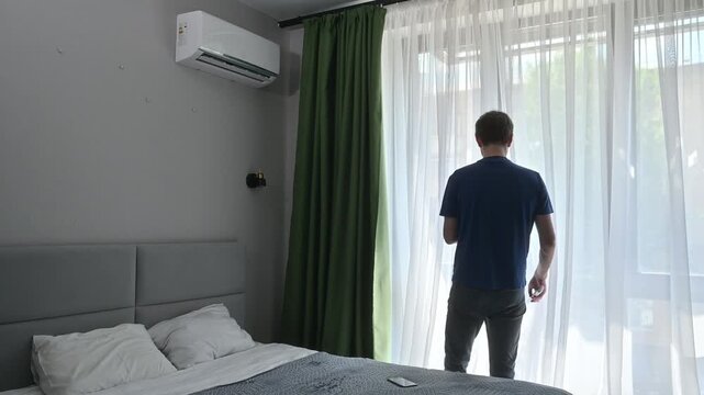 Man turns off the air conditioner with a remote control, then walks to the window and opens it