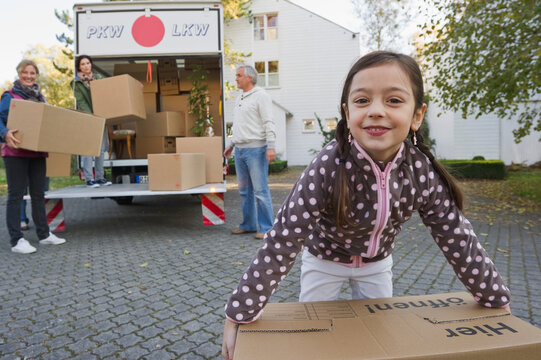Smiling girl carries a box while family loads moving truck outdoors