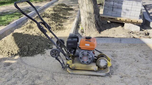 Vibratory plate compactor machine resting on sand base layer at construction site outdoors daytime. Equipment for compacting soil aggregate preparation for paving stone installation.
