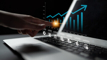 Fototapeta premium A close-up view of a hand interacting with a laptop screen showcasing a financial graph illustrating business growth trends and relevant icons in a dark atmosphere. Ledger