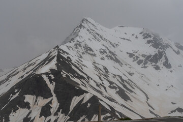 Obraz premium Picturesque Grossglockner High Alpine Road in Austria surrounded by rugged snowy peaks, green Alpine slopes and dramatic cloudy mountain scenery.