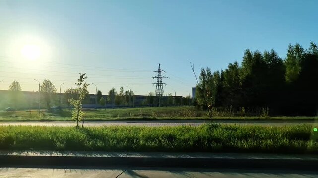 Roadside view from moving car passing urban industrial area with power lines and bright sun. Landscape includes green grass and trees along path of vehicle, clear sky background.