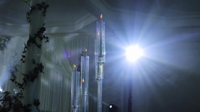 Tall glass candelabras featuring flickering flame electric candles, illuminated by a brilliant light creating a prominent lens flare, capturing an elegant indoor event atmosphere slow motion