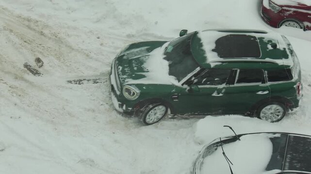 Green car stuck in deep snow drift with wheels spinning attempting to move on winter road. Vehicle bogged down after heavy snowfall, slow motion
