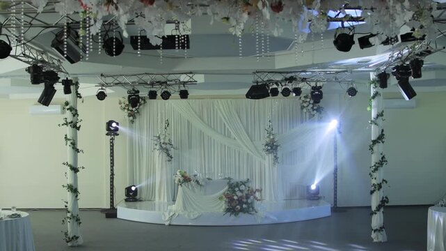Elaborate wedding reception stage decoration with elegant white drapes, luxurious floral arrangements, professional lighting setup in empty event hall prepared for celebration slow motion