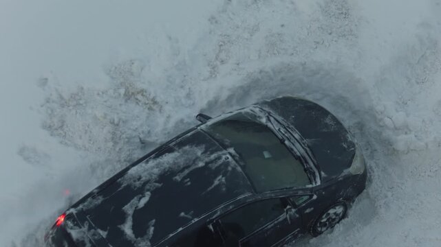 Aerial view of dark passenger car stuck in deep winter snow with wheels spinning, attempting to free itself from snowdrift.