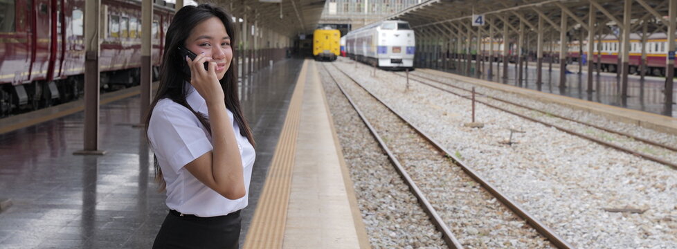 A radiant young Asian woman with long black hair stands confidently in a crisp white short-sleeved collared shirt and black skirt, smiling warmly at the camera while holding a phone to her ear