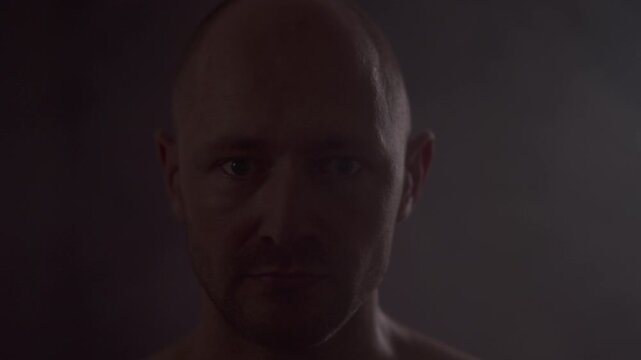 Intense man staring into camera then slapping his own head to snap out and stay focused. Bald male hitting his forehead, returning to serious expression in low key dark studio with subtle smoke.