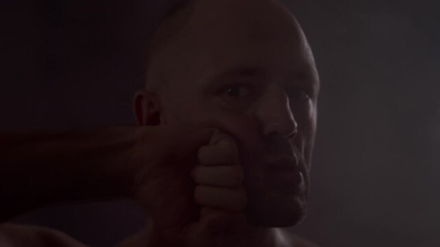A dark, atmospheric video: a close-up portrait of a bald man indoors, staring intensely at the camera, then slowly punching himself in the jaw.