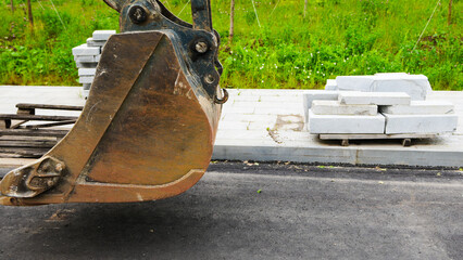 Large steel excavator bucket suspended above asphalt at a construction site with concrete paving materials in the background, showing heavy machinery and road construction equipment © belavinstock