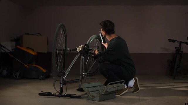 Young man crouching, holding a flashlight and working on the drivetrain of an upturned bicycle, with a toolbox beside him in a home garage