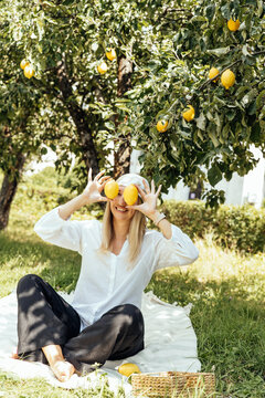 Smiling woman holding fresh lemons while sitting under a lemon tree in a sunny orchard