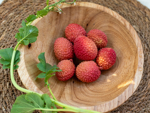 Ripe lychee fruits, Litchi chinensis, arranged in a natural wooden bowl with green foliage on a rustic woven surface. Warm food styling emphasizes tropical fruit texture, healthy nutrition and fresh e