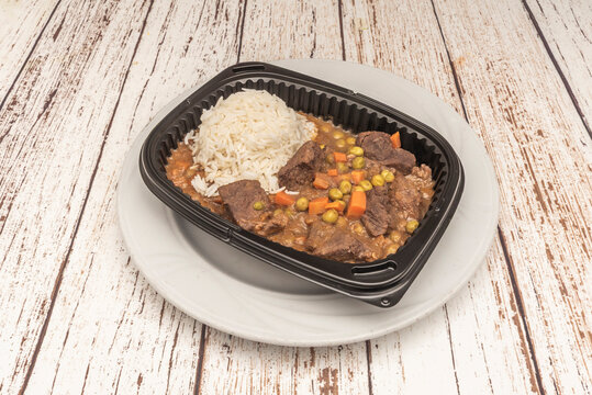 Beef stew with peas and carrots served alongside white rice in black takeout tray on plate.