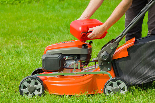 Man hands holding 5L red plastic canister and filling petrol in lawn mower tank on green grass at home yard in sunny summer day. Closeup. Side view.