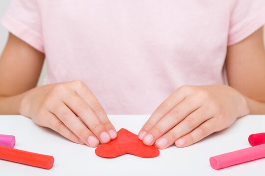 Little girl hands making and creating heart shape from red modeling clay on white table at home room. Closeup. Little child handcraft. Front view.