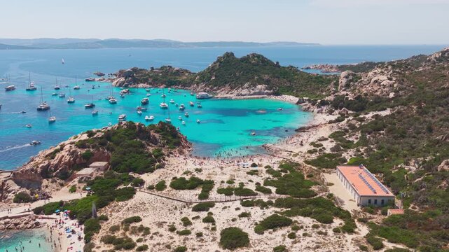 Wide aerial flying over luxury sailing yachts in turquoise blue water, with a beautiful sandy beach nestled in granite cliffs on the Isola di Spargi in the Maddalena Archipelago in Sardinia, Italy.