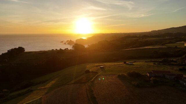 Drone flight above rural fields and scattered houses facing a bright sunset over the Asturian coast.